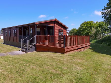 A wooden cabin with a fenced deck and steps on a lawn with trees in the background at Lodge 9- Lamorna in Tregolds near St Merryn