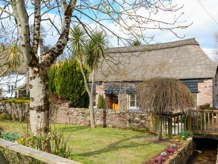 A thatched roof cottage with stone walls surrounded by trees and a small wooden bridge over a stream at Meadow Thatch in Cockington