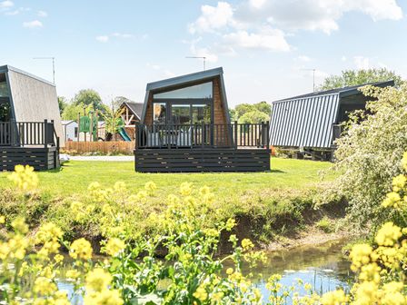 A grassy area with modern small cabins next to a small pond with yellow flowers and a playground in the background at Celestial Skylight - Billing Aquadrome in Billing Aquadrome Holiday Park