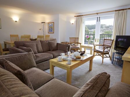A living room with brown sofas a wooden coffee table set with cups and plates two chairs by a window with curtains and a dining table with chairs at 10 Dartmouth House in Dartmouth