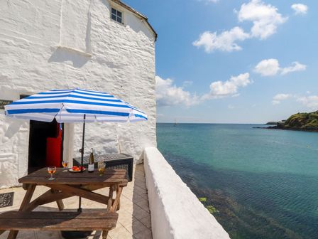 An outdoor patio with a wooden picnic table under a blue and white striped umbrella overlooking the sea at Rock Cottage in Portmellon near Mevagissey