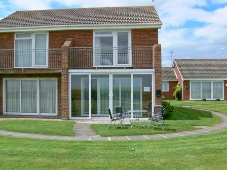 A two-story brick house with sliding glass doors on the ground floor and a balcony above with outdoor chairs and a table on a lawn at Seascape in Kessingland near Lowestoft