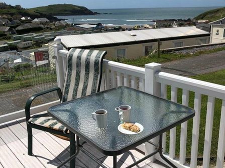 a balcony with a table, two cups, and a plate of food overlooking the sea at Challaborough Bay Holiday Park - Holiday Accommodation 11733 in Kingsbridge