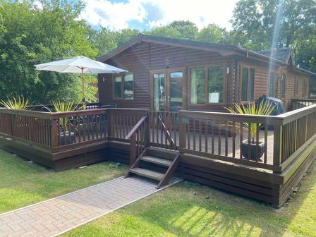 a wooden lodge with a deck surrounded by greenery at St Minver Holiday Park - Holiday Accommodation 15396 Wadebridge