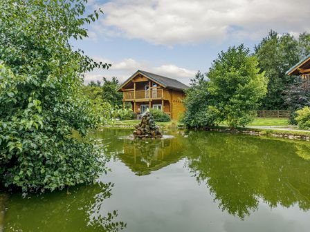 A house by a pond with trees and a fountain at Birch Waterside Spa in Carlton Miniott near Thirsk