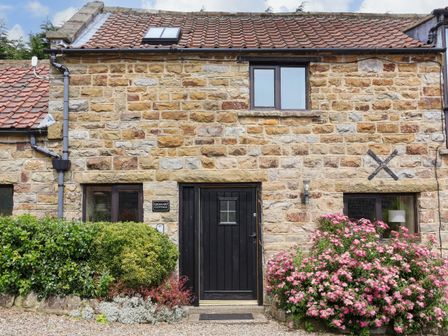A stone cottage with a black door and windows surrounded by green bushes and pink flowers at Granary Cottage in Staintondale