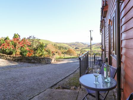 An outdoor patio with a glass table and chairs beside a wooden building overlooking a garden and hills at Beech Cottage in Llanidloes