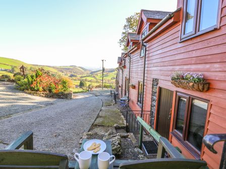 A view of a red wooden building with flower boxes a plate of biscuits and two mugs on a wooden railing overlooking a countryside landscape at Oak Cottage in Llanidloes