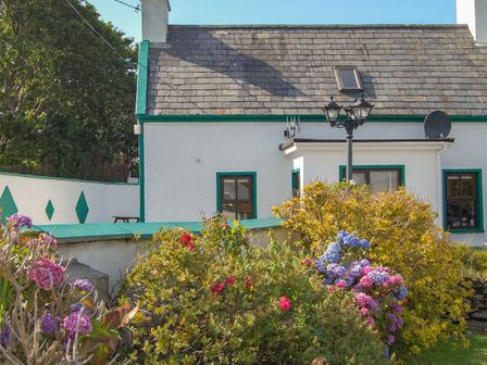 A white house with green trim and a slate roof with flowers and shrubs in the garden at Nana's House in Allihies County Cork