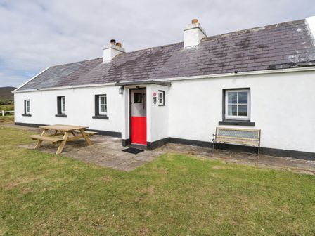 A white single-story cottage with a red door wooden picnic table and a bench outside at Sound Cottage in Achill Island County Mayo