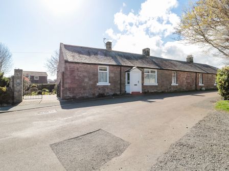 A stone cottage with a white door and windows beside a road and a gated entrance with a garden behind at Lyndhurst Cottage in Beadnell