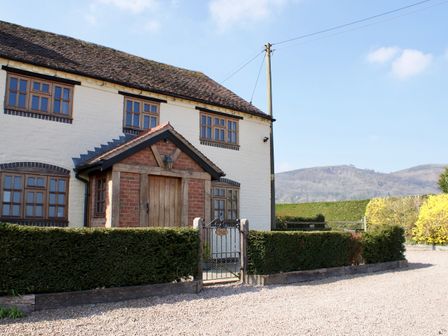 A white brick house with multiple windows and a wooden door behind a hedge and gate at Yew Tree Cottage in Little Malvern near Malvern
