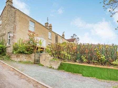 A stone cottage with plants and a hedge along a gravel driveway at Hillside Cottage in Peasedown St John near Bath