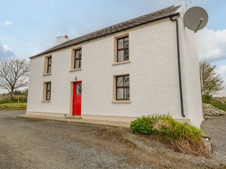 A white two-story house with a red door and several windows with brown frames with gravel driveway and some greenery at Mary Kate's Cottage in Narin County Donegal