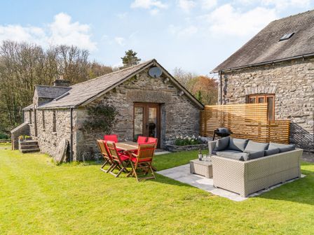 Outdoor patio area with wooden table and chairs with red cushions and a woven sofa set on grass at Moresdale Bank Cottage in Lambrigg near Sedbergh and Kendal