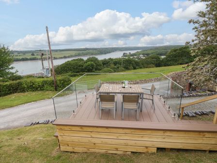 A wooden deck with a table and four chairs overlooking a river and green landscape at Penrallt Cottage in Y Felinheli near Caernarfon