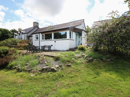Exterior of a white cottage with a patio set on a stone terrace surrounded by grass and shrubs at Morfa Isaf Cottage in Llangrannog