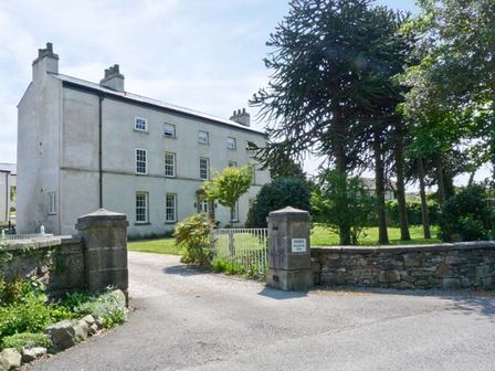 A large three-story house with multiple windows and chimneys next to a gated driveway with stone pillars and surrounding trees at 2 Cark House in Cark in Cartmel