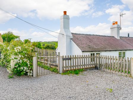 A white cottage with chimneys and a wooden fence with a metal gate surrounded by gravel and greenery at Quarry Cottage in Trelogan near Llanasa