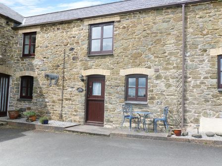 A stone building facade with two windows a wooden door metal wall art and a small table with two chairs at The Old Granary in Polyphant near Launceston