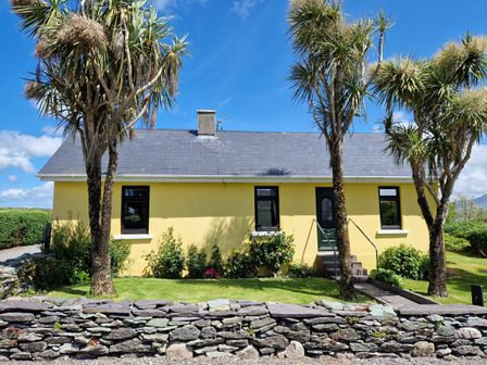 A yellow house with a gray roof surrounded by grass and trees behind a stone wall at Kates Cottage in Knightstown Valentia Island County Kerry