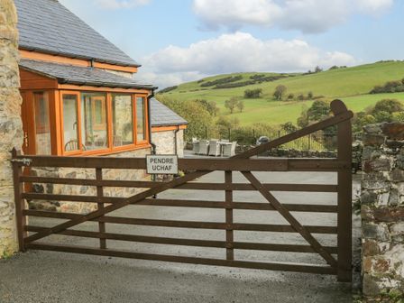 A wooden gate with a sign reading Pendre Uchaf next to a stone building and outdoor seating with hills in the background at Pendre Uchaf in Melin-y-Wig near Ruthin