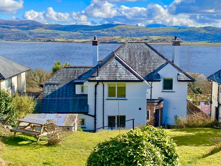 A house with a grey slate roof and white walls overlooking a large body of water with hills in the background at Bryn Eithin in Borth-y-Gest near Porthmadog