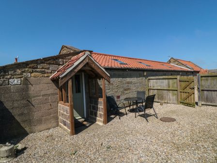 A stone building with a tiled roof and a small porch next to a gravel courtyard with a table and chairs at The Goat Shed in High Normanby near Robin Hood's Bay