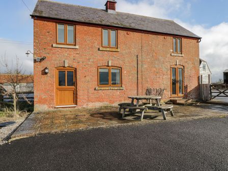 Exterior view of a red brick house with wooden framed windows and doors and a wooden picnic table on a paved area in front at Stables Cottage in Bowerhill near Melksham