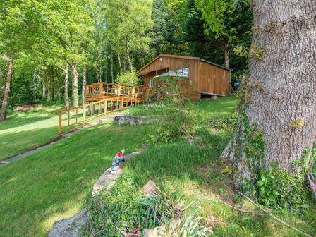 A wooden cabin with a deck and umbrella surrounded by grass and trees at Bluebell Cottage in Maenan near Llanrwst