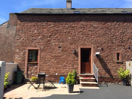 A stone building exterior with a door, windows, potted plants, a table with chairs, and a small blue chair at The Old Bothy in Bolton Low Houses near Wigton