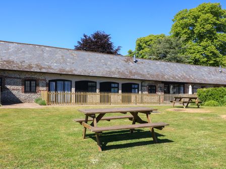 A grassy outdoor area with wooden picnic benches and a building with a tiled roof and fenced patio at The Old Stables in Winterborne Stickland