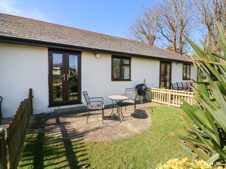 An outdoor patio area with a small table and two chairs and a barbecue grill outside a white building with brown doors and windows at Honeysuckle Cottage in Bude