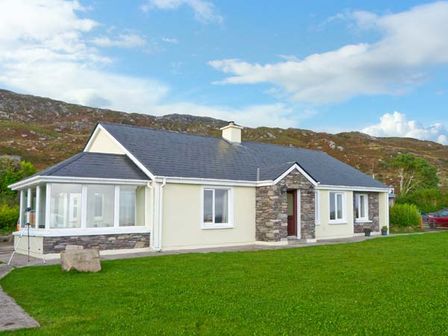 A single story house with stone and cream walls and a glass porch on a grassy lawn at Kerry Way Cottage Castlecove County Kerry