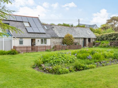 A garden area with a flower bed and grass in front of a building with solar panels and wooden fences at Stable Cottage in Ilfracombe