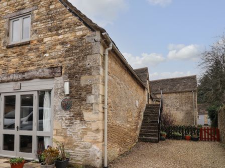 Exterior of a stone barn with a glass door and stone steps leading to an upper level with potted plants and a red gate at Greyhound Barn in Barnsley near Cirencester