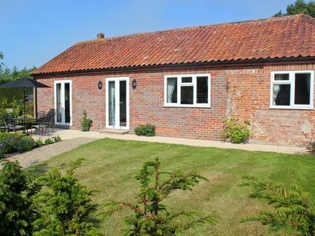 A brick house with a tiled roof and windows with white frames a lawn and small bushes in front with an outdoor table and chairs under a black umbrella at Moat Farm Cottage in Wood Dalling