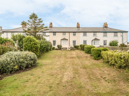 A row of terraced houses with chimneys and a grassy garden area with bushes and a picnic table at Coastguard Cottage in Low Newton-by-the-Sea near Beadnell