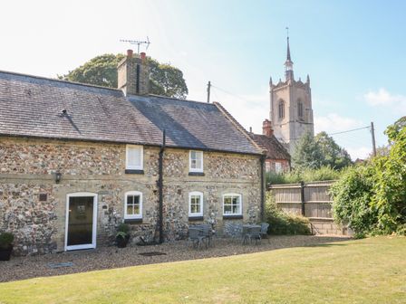 A cottage with stone walls and a slate roof with outdoor seating and a church tower in the background at Manor Farm Cottage in Swaffham