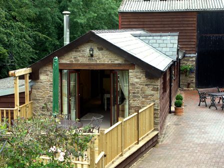 An outdoor view of a stone and wood cottage with open glass doors a wooden fence and outdoor metal furniture at Cwm Derw Cottage Llanafan Fawr near Builth Wells