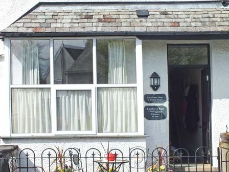 The exterior of a white house with a tiled roof a large window a black door small garden with flowers and a small table with chairs at Swallows Nest in Coniston