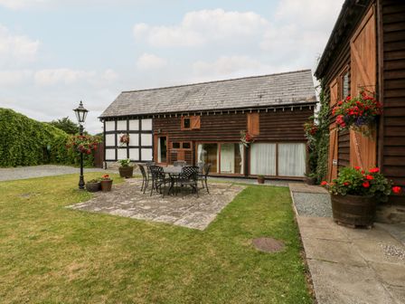 An outdoor patio area with a metal table and chairs surrounded by a grass lawn and wooden buildings with flower pots at Stable End in Luntley near Pembridge
