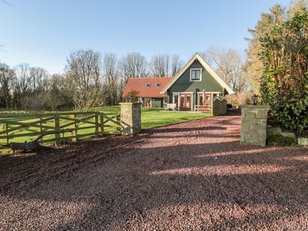 A driveway with gravel leading to a house with a red roof and green siding surrounded by a lawn and trees at Apple Tree Lodge Swarland near Alnwick
