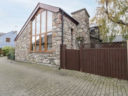 A stone exterior building with large windows and a brown wooden fence next to a tree at Ddol Helyg Barn in Llanrug
