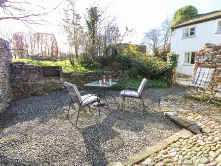 A garden patio area with stone walls two cushioned chairs and a table with wine bottle and glasses at Cherry Tree Cottage in Soulby near Kirkby Stephen