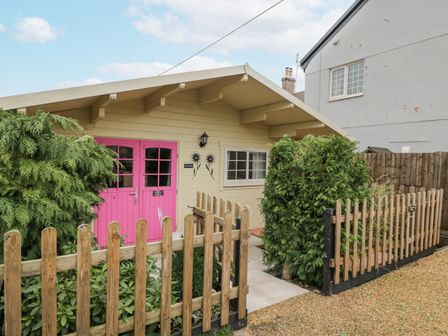 An exterior view of a small wooden cabin with pink double doors surrounded by greenery and a wooden fence at The Log Cabin in Bushton near Royal Wootton Bassett