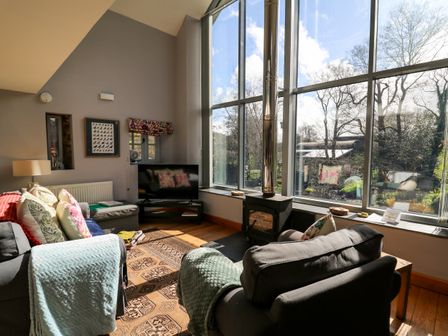 A living room with armchairs a wall-mounted tv and a wood-burning stove by large windows at The Barn at Glanoer in Hundred House near Builth Wells