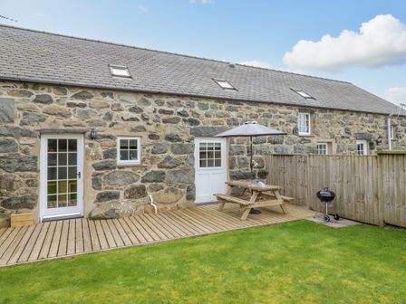 A stone exterior of a house with a wooden deck a picnic table with an umbrella a charcoal grill and a grassy yard at Ysgybor Yd in Chwilog near Criccieth