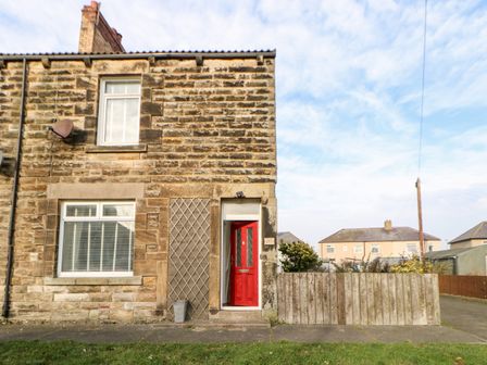 The front of a stone house with a red door and two windows beside a wooden fence at Lightkeeper House in Amble-by-the-Sea