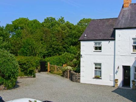 A white house with a dark roof and two windows next to a gravel driveway with green trees and bushes in the background at Carreg Rhys in Malltraeth near Newborough